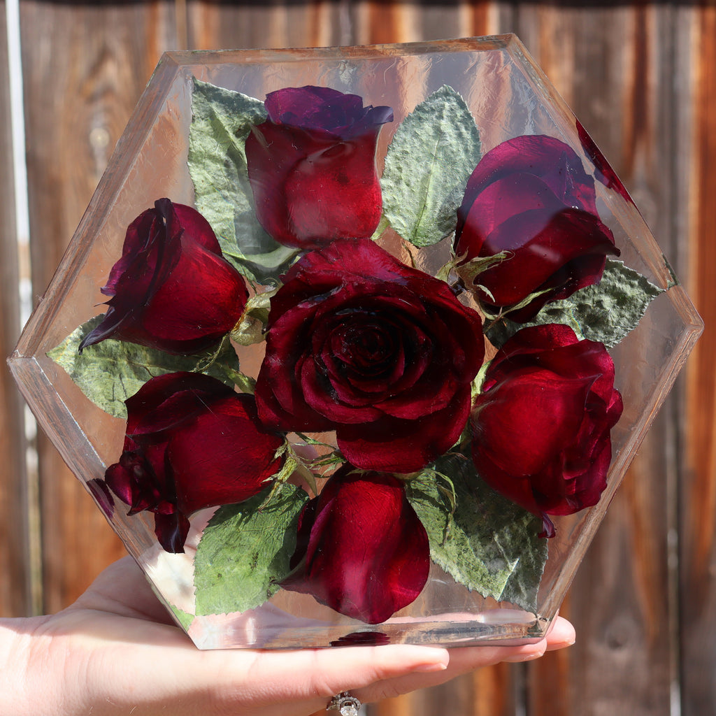 Hexagonal resin block containing preserved red roses held by a hand against a wooden fence background